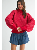A woman wearing a bright red knit sweater with a textured geometric pattern and voluminous balloon sleeves, paired with blue denim shorts, against a light gray studio background.